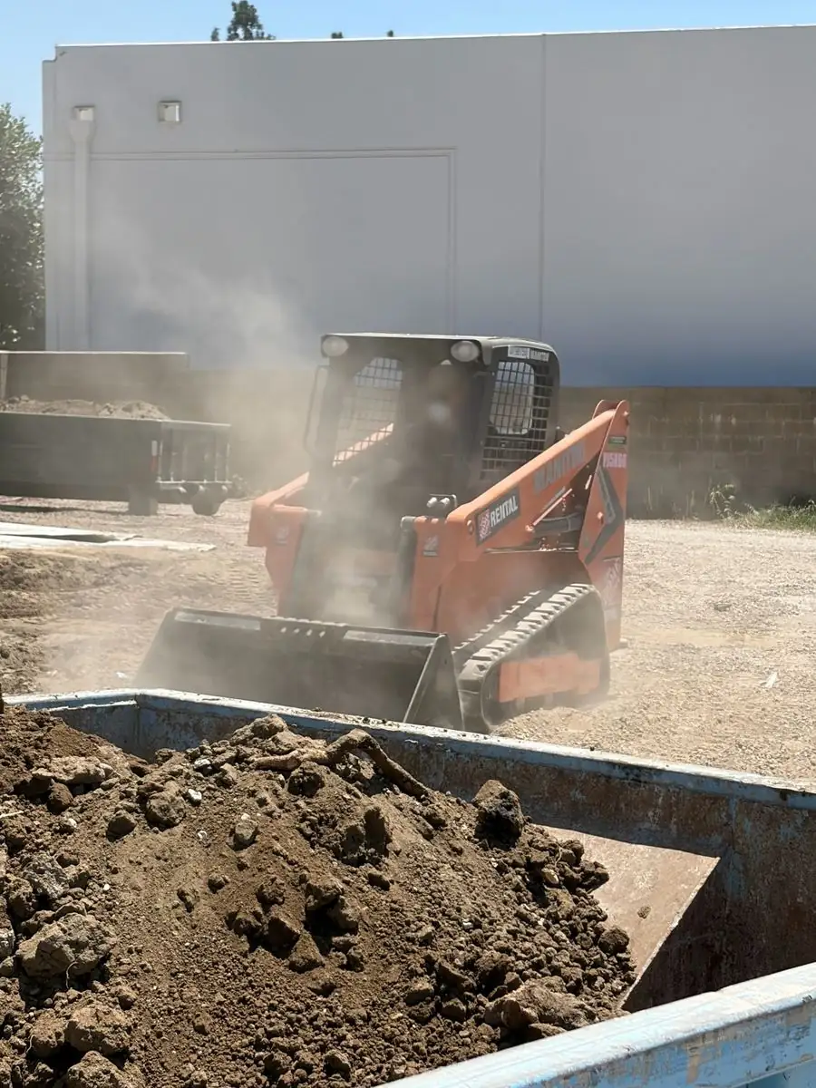 Skid steer kicking up dust as it loads dirt into a roll-off bin on a job site